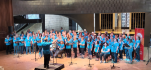 Long shot people in blue singing onstage with conductor